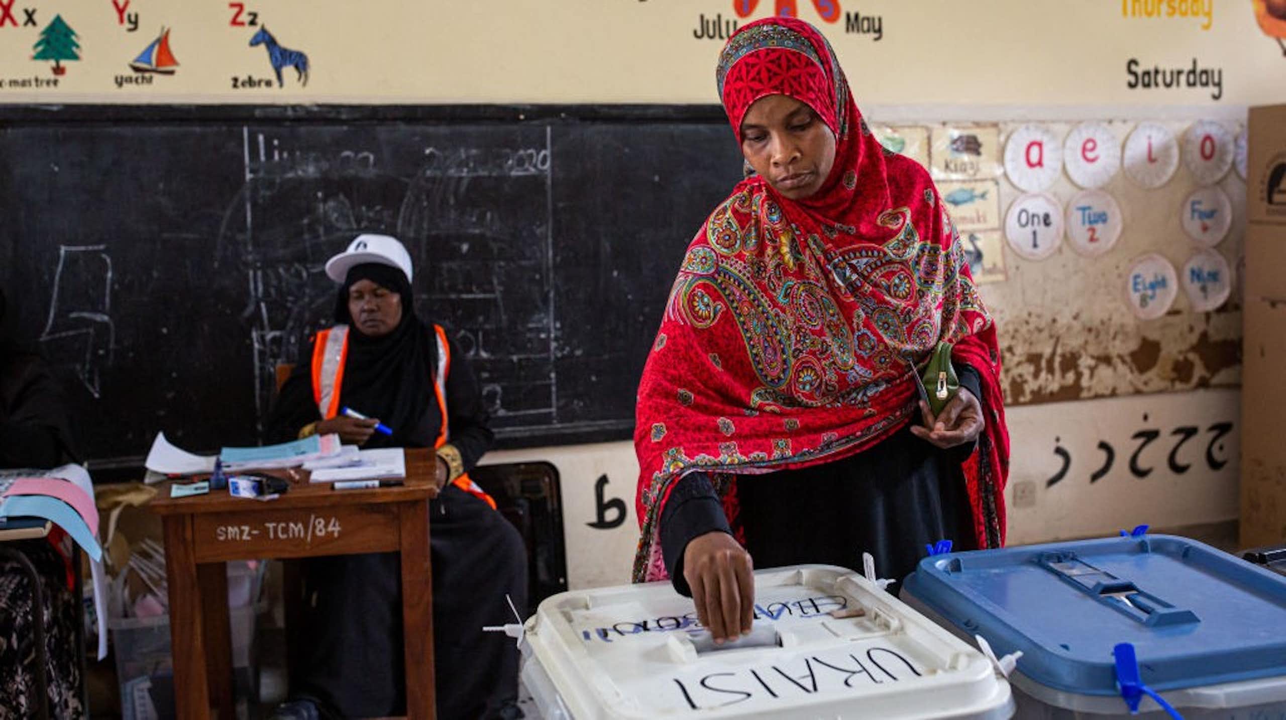 A woman wearing a headscarf places her ballot into a box in a school classroom.