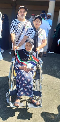 An elderly woman in a wheelchair, holding a South African flag and draped in the colours of the Palestinian flag, is flanked by a man and a woman