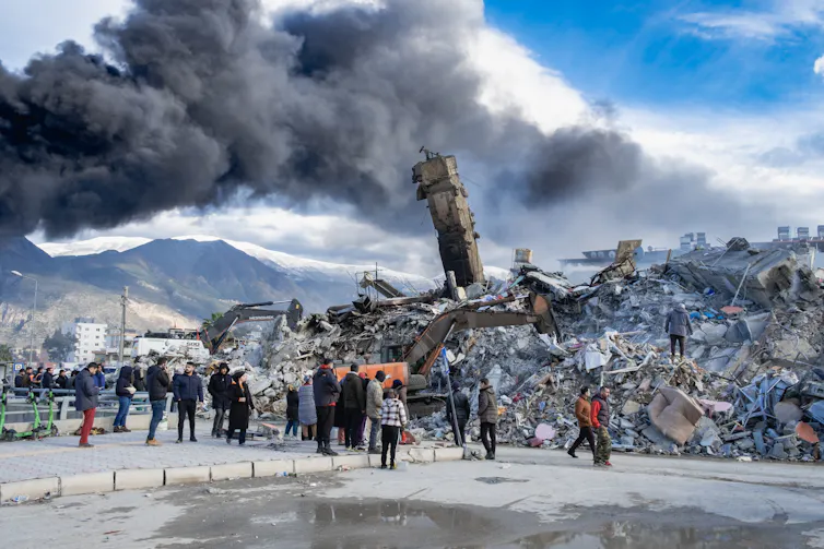 A group of people standing in front of crumbled buildings with a cloud of smoke overhead.