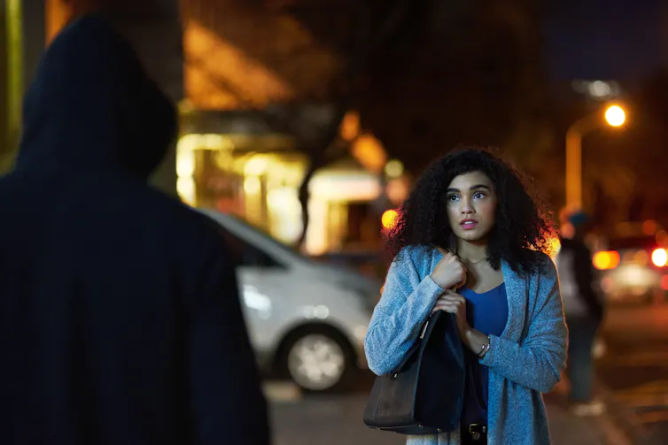 A young woman on a street at night looks fearful, a hooded figure is in the foreground appearing to confront her