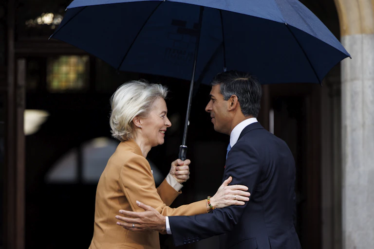 Ursula von der Leyen and Rishi Sunak greet each other with smiles under a large blue umbrella