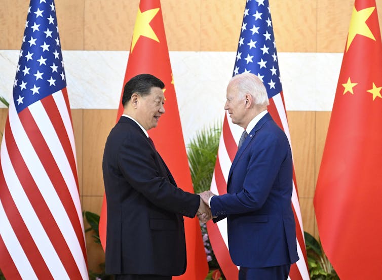 Xi Jinping and Joe Biden shake hands in front of a row of Chinese and American flags.