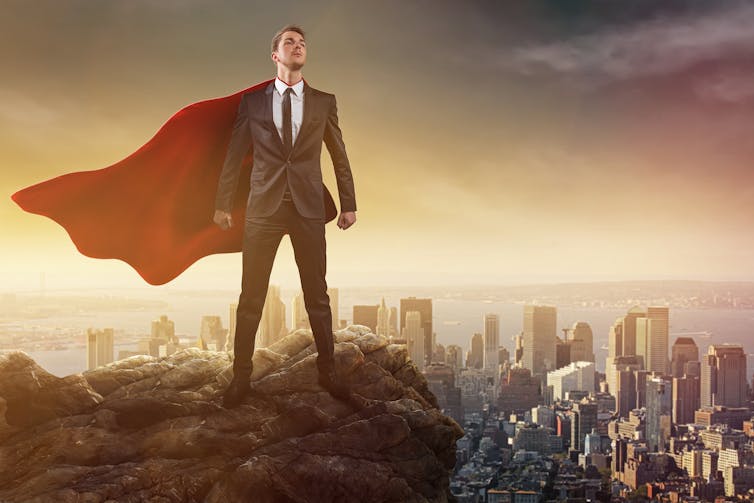 Man wearing cape and business suit perched above cityscape.