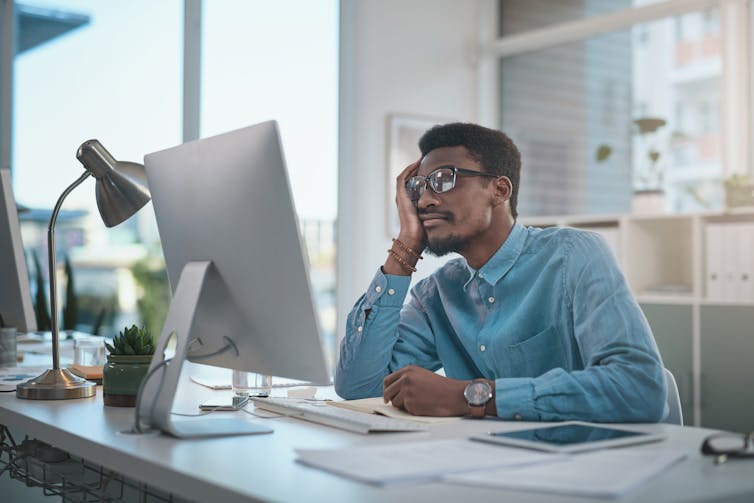 A bored man at desk leans his head in his hand while looking at a computer.