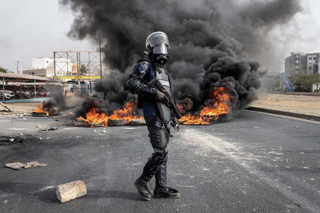Armed man in police gear, standing in front of burning tyres on a road.