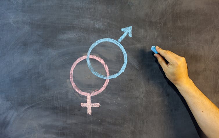 A hand is seen drawing on a black board next to symbols of the male and female genders.