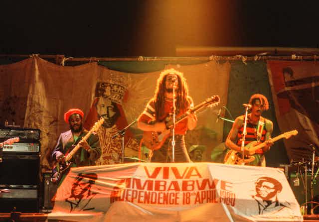 Un hombre con rastas canta y toca la guitarra delante de un cartel en el que se lee "Viva Zimbabue".
