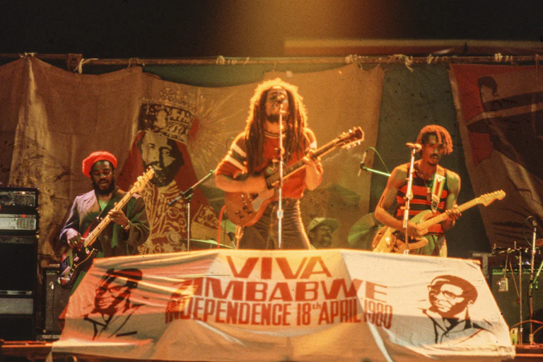 Un hombre con rastas canta y toca la guitarra delante de un cartel en el que se lee "Viva Zimbabue".