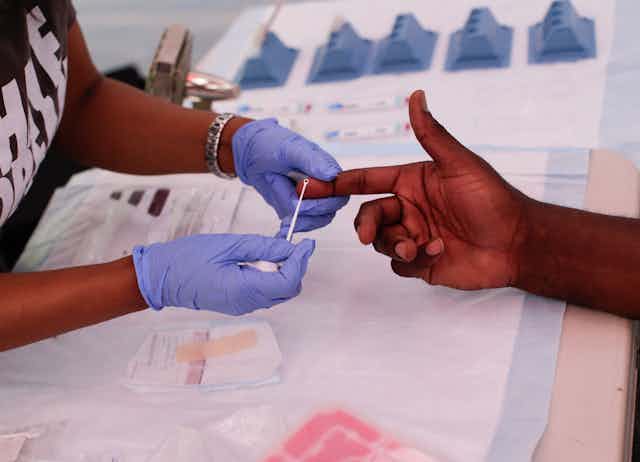 A person wearing gloves is holding a Black man's finger as they extract a sample of blood.