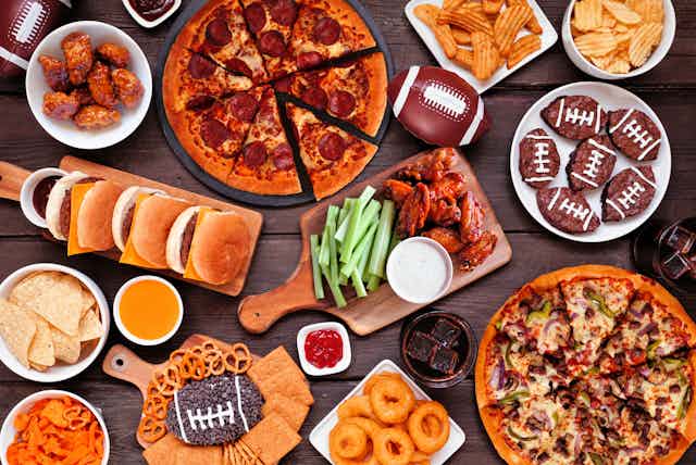 An overhead view of a table filled with all kinds of food, including pizza, chips and dip, cookies decorated like footballs, pretzels and onion rings.