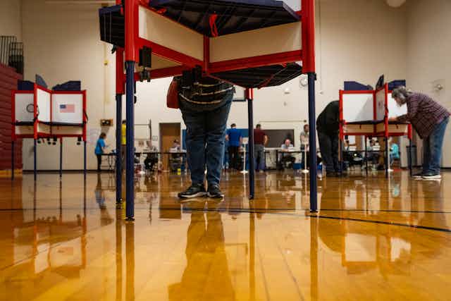 People stand behind white and red voting divisions in what looks like a school gymnasium, with a shiny brown floor. 