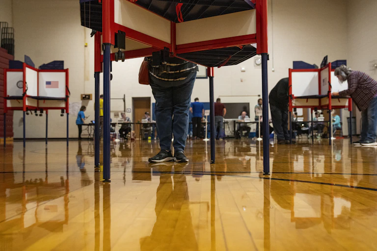 People stand behind white and red voting divisions in what looks like a school gymnasium, with a shiny brown floor.
