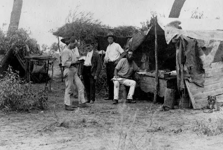 A black and white photo of a group of men standing in a bush campsite.
