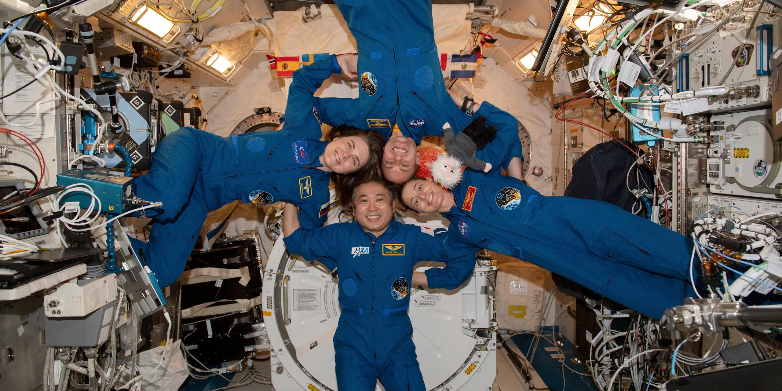 Four astronauts in jumpsuits float with locked arms in the International Space Station.