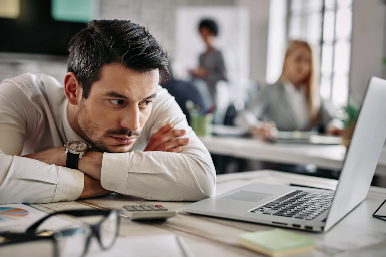 A man rests his chin on his crossed arms and stares morosely at an open laptop screen