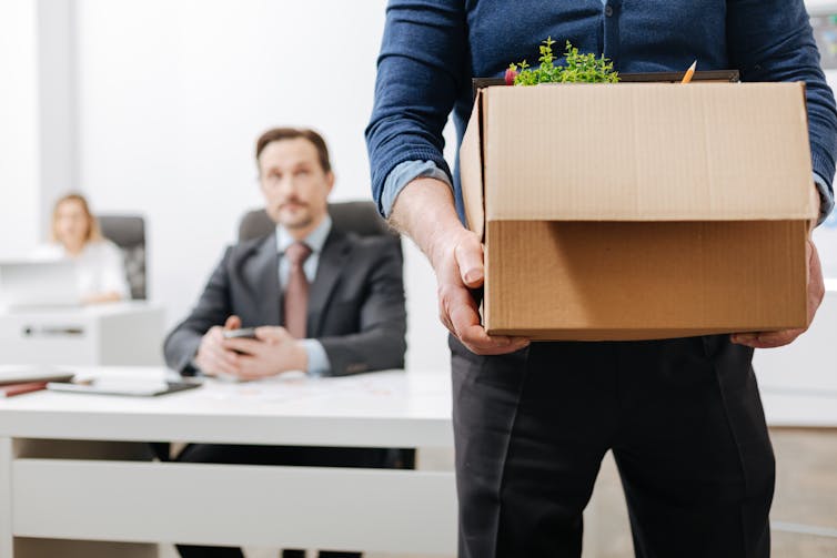 A man walks toward the camera holding a cardboard box with a plant peeking out the top. Behind him, a man in a suit sits at a desk with his hands folded in front of him.