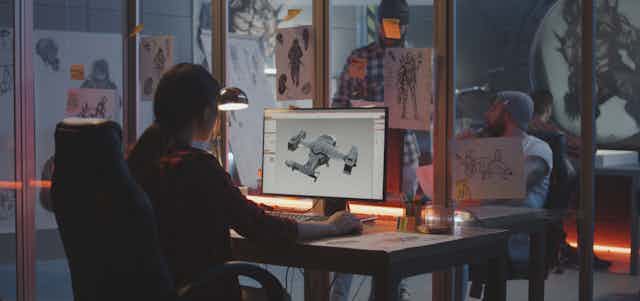 A woman in an office works on a computer. Drawings are on the walls. Two men are talking in the background