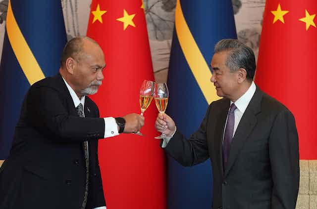 Two men standing in front of the China and Nauru flags clinking their drinks glasses together.
