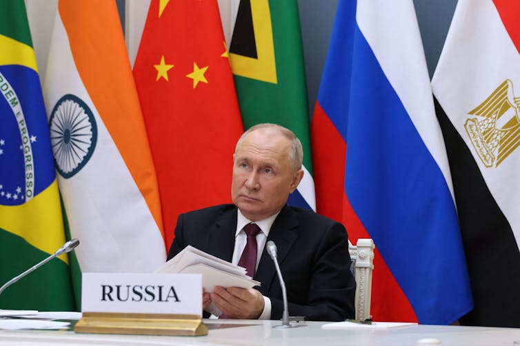 A man in a suit shuffles papers behind a sign reading Russia. Behind him are various flags.