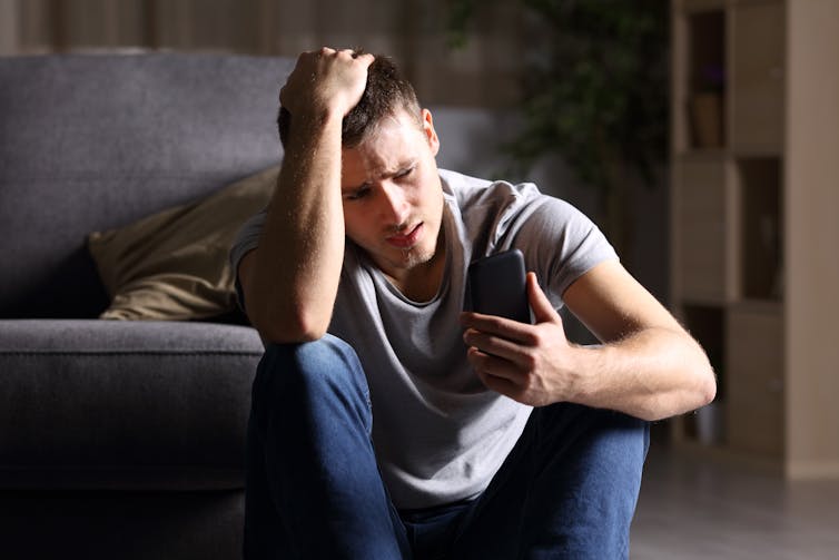 A young man sitting on the floor against a couch, looking at his phone with a disappointed and confused look on his face