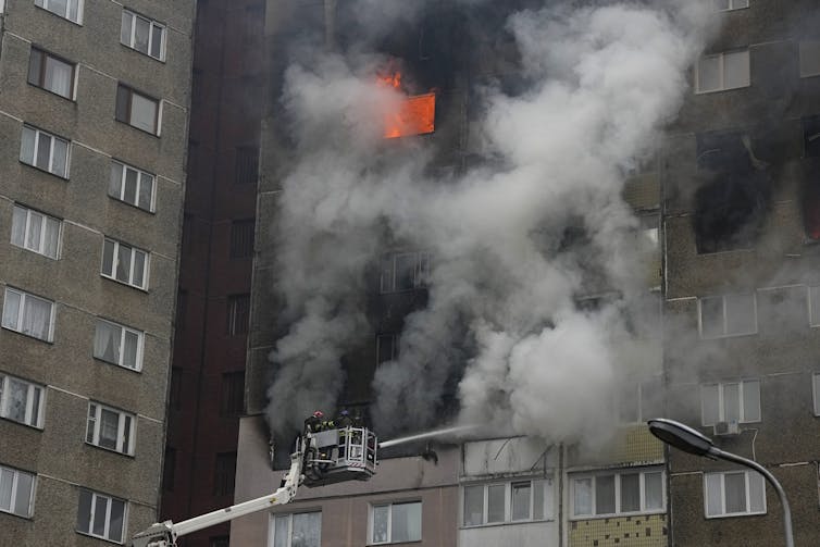 A fire engulfs part of an apartment building as firefighters in a cherry picker point a hose at it.