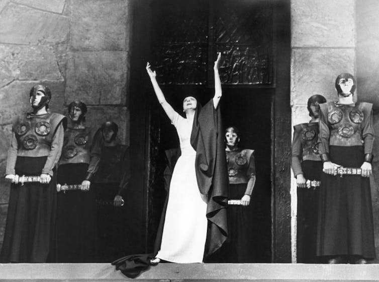 A woman in a white dress and black shawl throws her arms up dramatically in front of stern-looking soldiers.