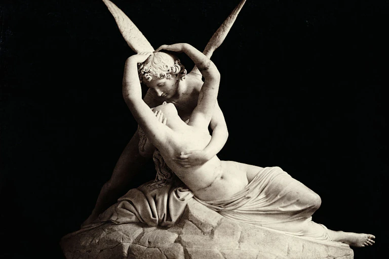 A white statue of a winged figure with curly hair embracing a reclining woman whose legs are mostly covered with a sheet.