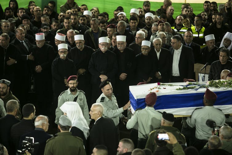 A large group of men, some of them wearing dark jackets and white hats, surrounding a coffin that has an Israeli flag draped over it.
