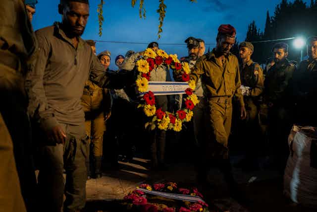 A group of men wearing army green hold a flower wreath that has writing in Hebrew in the center of it. 