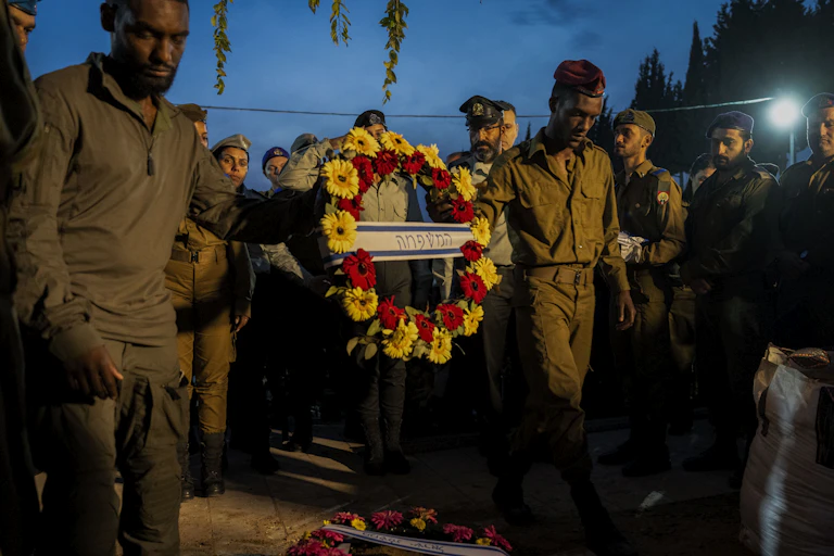 A group of men wearing army green hold a flower wreath that has writing in Hebrew in the center of it.