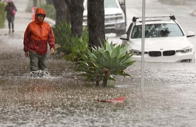 A man walks through knee-high water on a sidewalk next to a car parked at the curb with water over its wheels.