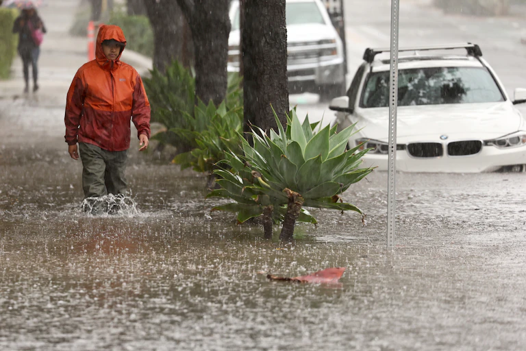 A man walks through knee-high water on a sidewalk next to a car parked at the curb with water over its wheels.