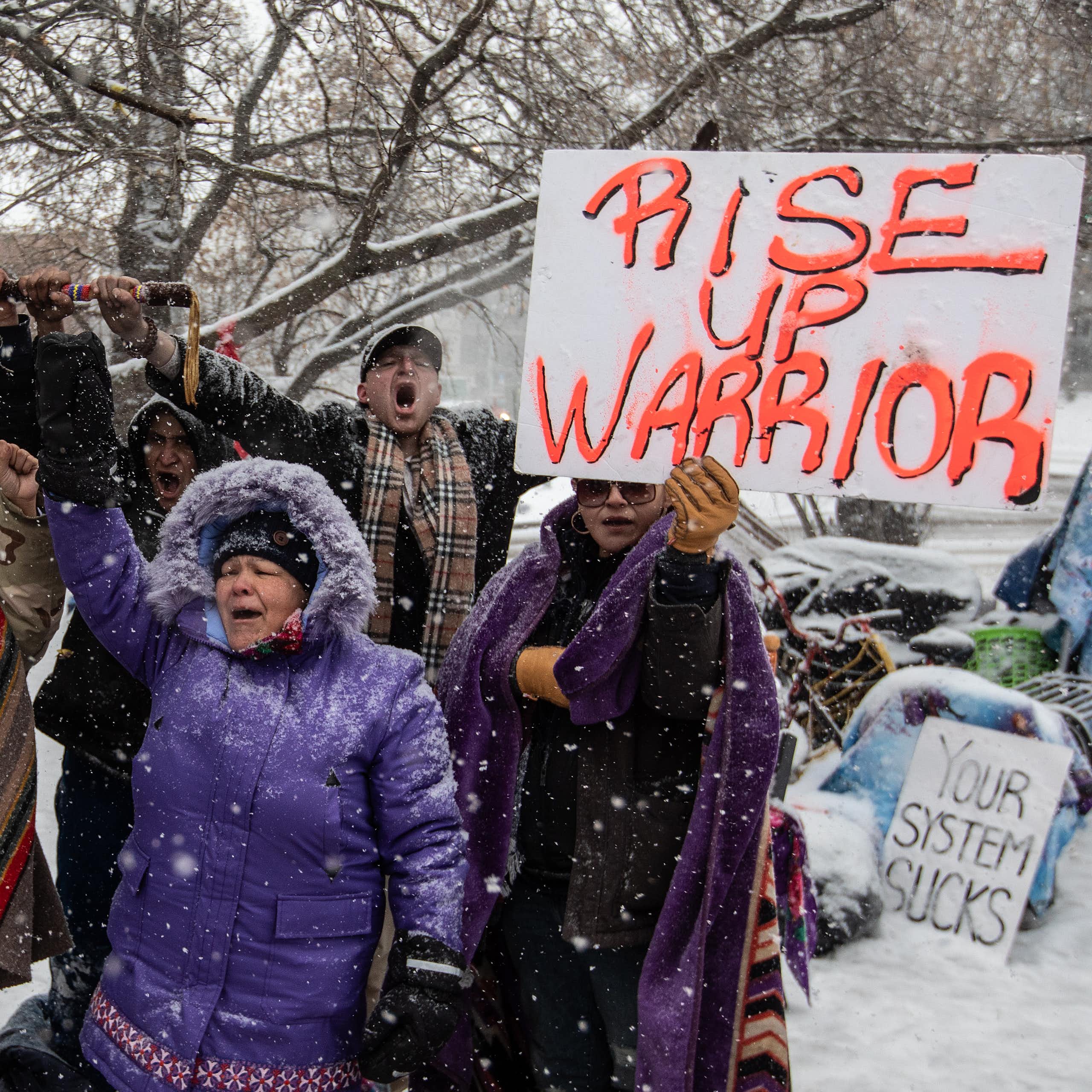 People in parkas protest in the snow, one carrying a sign that reads Rise Up Warrior.