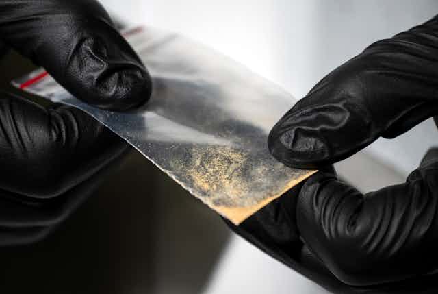 Gloved hands holding small clear packet of brown powder substance.
