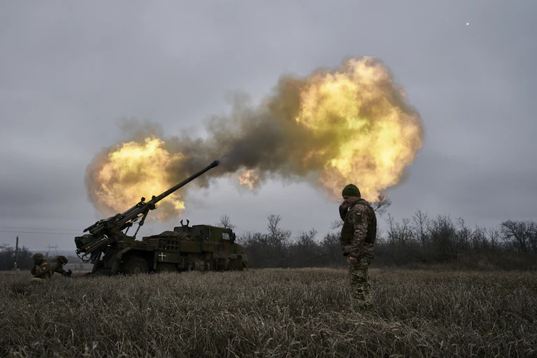 Soldier standing in field covering ears as another solder fires an artillery shell, a cloud of burning smoke rising in the sky