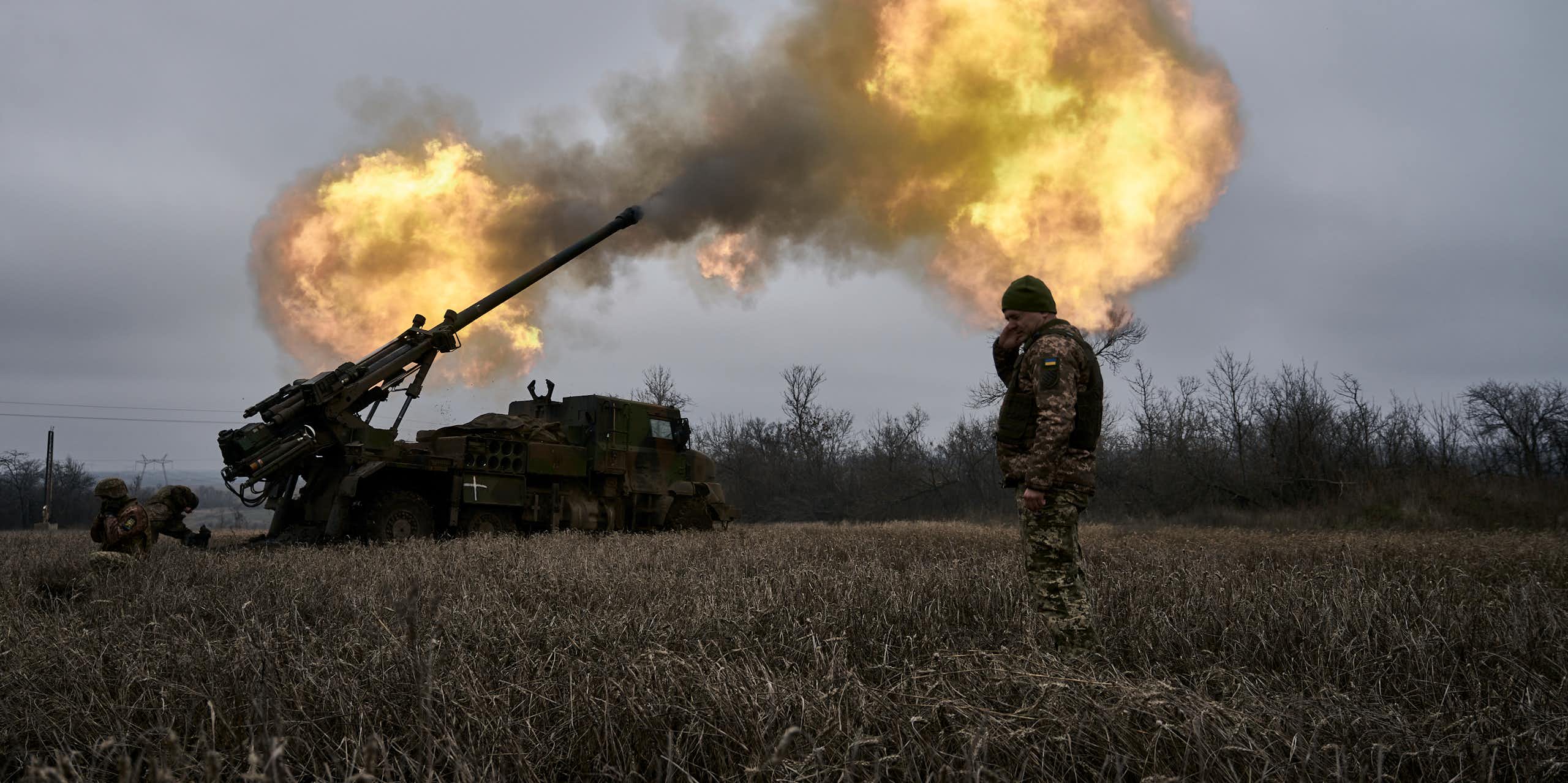 Soldier standing in field covering ears as another solder fires an artillery shell, a cloud of burning smoke rising in the sky