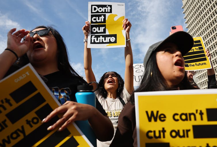 Three female protestors shout, while one holds a sign reading 'Don't cut our future.'