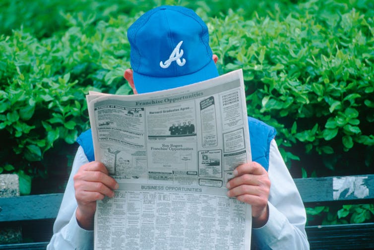 Man wearing blue hat sits on a bench reading a newspaper.