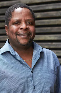 An African man in a blue shirt smiles at the camera.