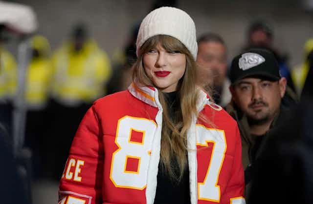 Taylor Swift smiling and wearing a jacket in the style of her boyfriend Travis Kelce's NFL jersey