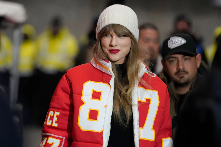 Taylor Swift smiling and wearing a jacket in the style of her boyfriend Travis Kelce's NFL jersey