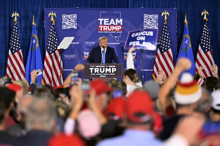 A man on at a lectern, flanked by American flags, talking to a crowd of people, many wearing red hats.