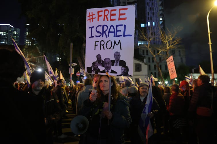 Crowd of protestors at night in a city, one holds up sign saying '#FREE ISRAEL FROM' with pictures of Netanyahu and other ministers.