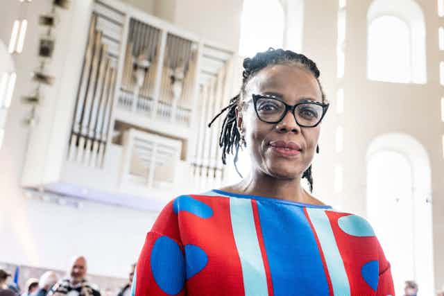 An African woman with glasses and greying dreadlocks smiles wryly at camera wearing colourful jersey and standing in a large hall with grand windows.