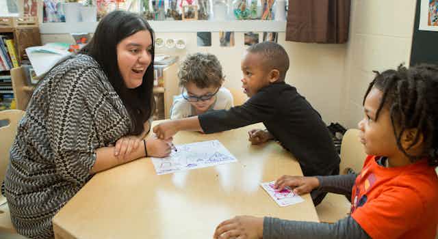 An adult seen at a table with children holding markers and paper.