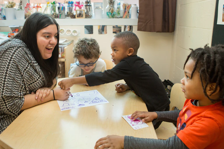 An adult seen at a table with children holding markers and paper.