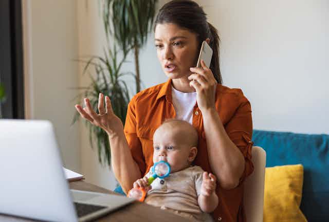 Une jeune femme parle au téléphone portable tout en regardant un ordinateur portable ouvert. Un bébé est assis sur ses genoux et mâchonne un jouet.