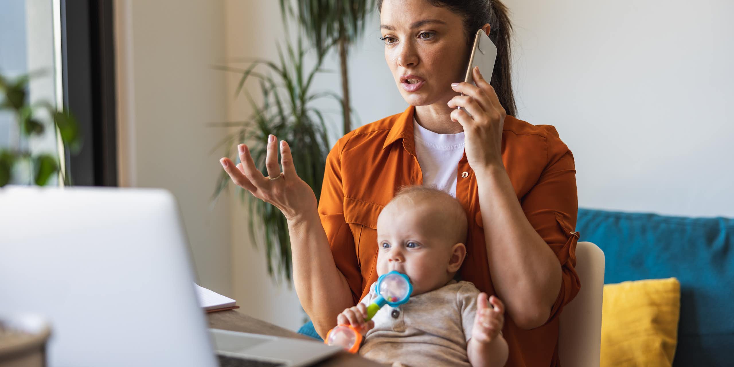 Une jeune femme parle au téléphone portable tout en regardant un ordinateur portable ouvert. Un bébé est assis sur ses genoux et mâchonne un jouet.