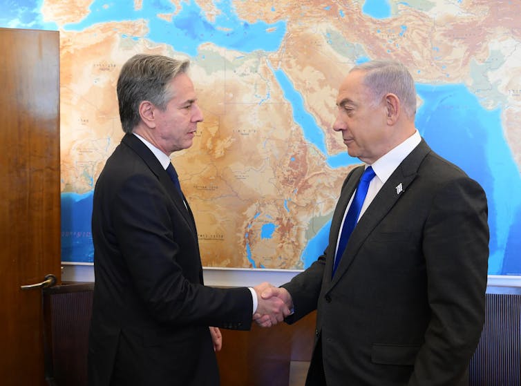 US secretary of state Antony Blinken shakes hands with Benjamin Netanyahu in front of a large map of the world.