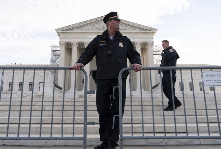 A police officer standing behind a barricade and in front of a large, white columned building.